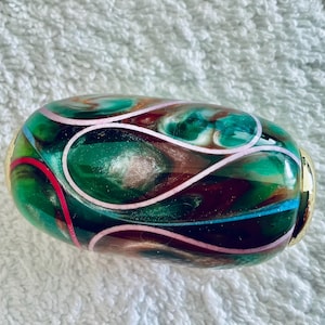 May include: An oval-shaped glass bead featuring a swirling pattern of deep green, dark red, and metallic bronze tones. The surface is decorated with thin, light pink lines that loop across the body, finished with polished gold-tone metal end caps.