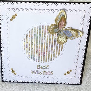 May include: A handmade greeting card with a white scalloped edge and a black border. The card features a gold and pastel butterfly, a striped circle, and the words "Best Wishes". Gold embellishments are placed around the card.