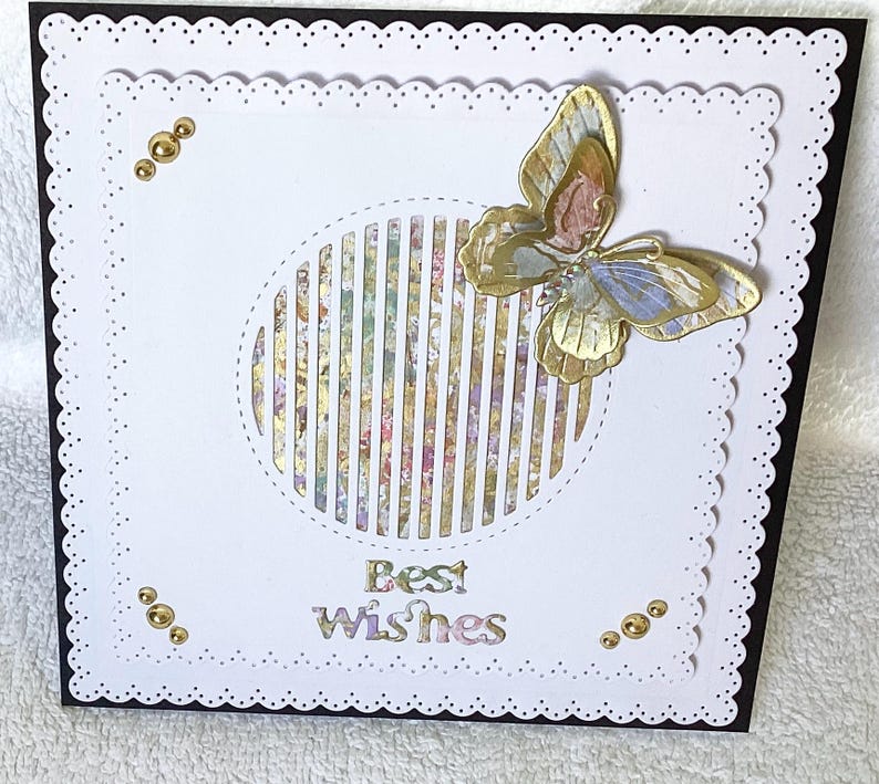 May include: A handmade greeting card with a white scalloped edge and a black border. The card features a gold and pastel butterfly, a striped circle, and the words "Best Wishes". Gold embellishments are placed around the card.