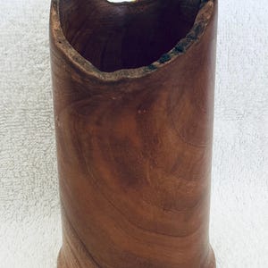 May include: A cylindrical brown wooden vessel with a natural, uneven, and textured rim. The wood grain is visible throughout the body. A yellow ruler is positioned behind the vessel, showing a height measurement of approximately 6 inches.