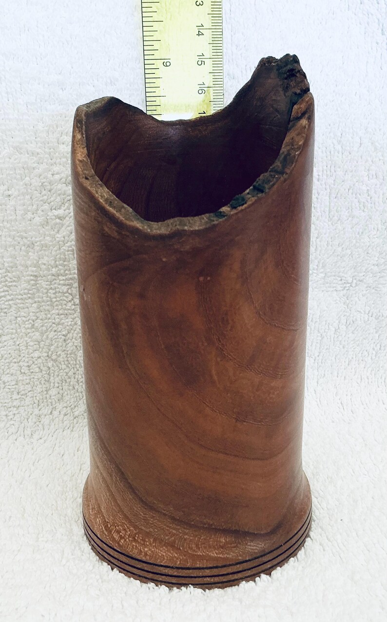 May include: A cylindrical brown wooden vessel with a natural, uneven, and textured rim. The wood grain is visible throughout the body. A yellow ruler is positioned behind the vessel, showing a height measurement of approximately 6 inches.