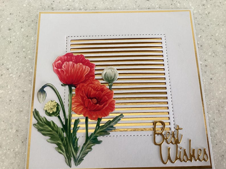 May include: A white greeting card with a gold border and accents. The card features a floral design with red poppies and green leaves. The words "Best Wishes" are in gold script.