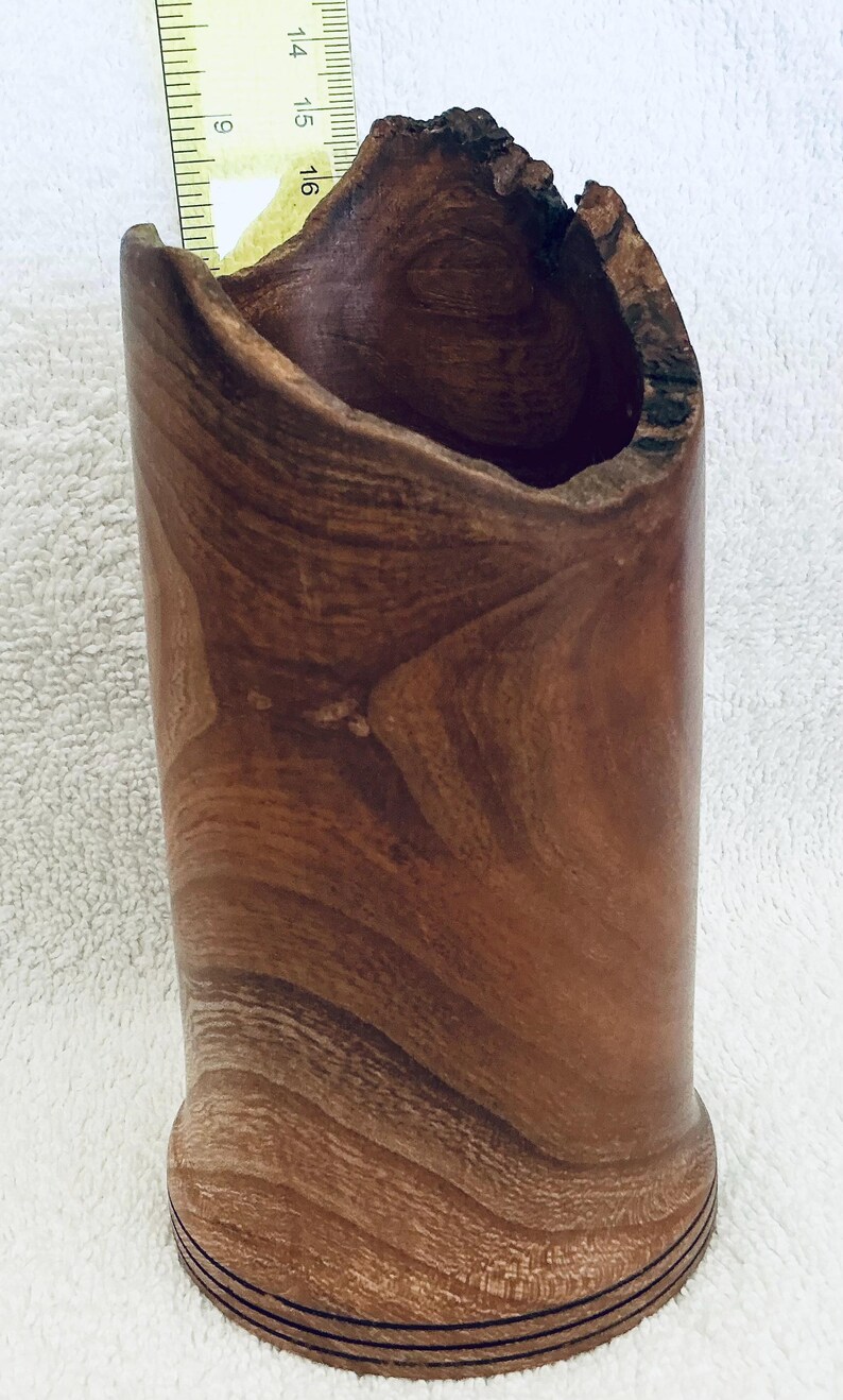 May include: A cylindrical wooden vessel with a natural live edge rim and visible wood grain patterns. A yellow ruler is positioned inside the opening, indicating a height measurement of approximately 6.5 inches from the base to the highest point.