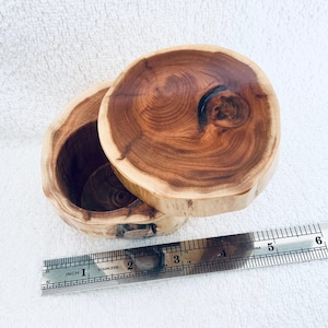 May include: A round wooden box with a natural live edge and a removable lid, featuring visible wood grain and a dark knot. A metal ruler is placed in front of the box, showing a diameter of approximately 9 cm for the lid and base.