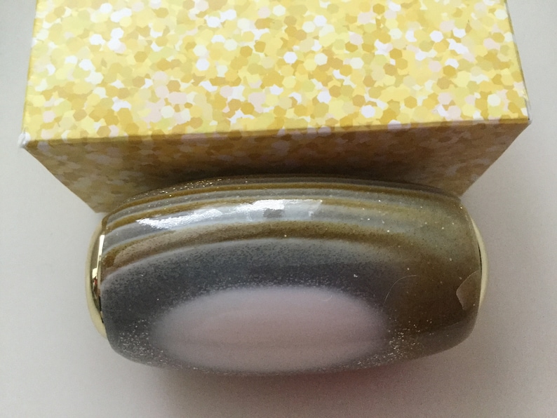 May include: A round, gray and gold bottle stopper with a pink center. The bottle stopper has a textured, speckled surface.