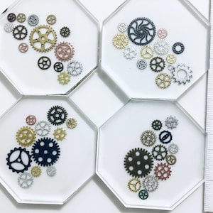 Puede incluir: Cuatro posavasos octogonales con fondo blanco, cada uno con una disposición de engranajes metálicos de varios tamaños en oro, plata, bronce y negro. Los engranajes están incrustados en resina transparente.