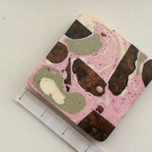 May include: A square, decorative object with a pink, white, and green marbled design. Dark brown wood pieces are inlaid throughout the surface. A ruler is placed next to the object, showing measurements in inches.