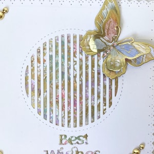 May include: A white greeting card with a decorative butterfly and the words "Best Wishes". The butterfly has gold, pink, and blue accents. Gold embellishments are placed around the card's edges.