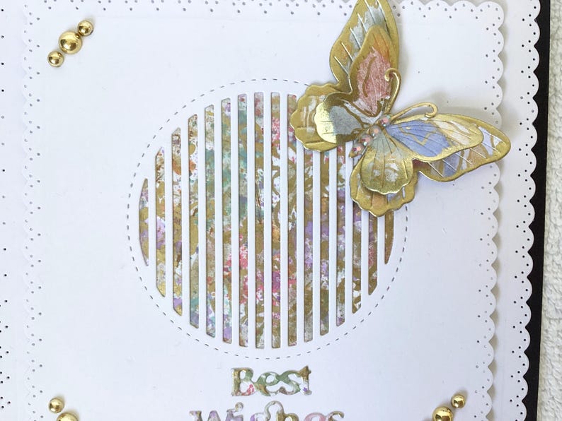 May include: A white greeting card with a decorative butterfly and the words "Best Wishes". The butterfly has gold, pink, and blue accents. Gold embellishments are placed around the card's edges.