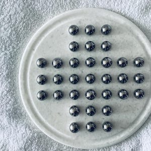 Peut inclure: Un jeu de solitaire en marbre avec 32 billes argentées brillantes disposées sur un plateau circulaire. Le plateau présente un motif en forme de croix pour les billes. Le jeu est posé sur une surface blanche texturée.