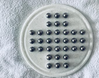 May include: A marble solitaire game featuring 32 shiny, silver marbles on a circular marble board. The board has a cross-shaped arrangement of holes for the marbles. The game is set on a white, textured surface.