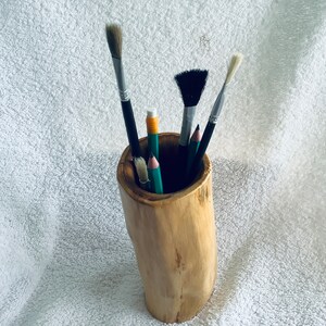 May include: A wooden pencil holder with a natural bark finish, filled with various paintbrushes and pencils. The holder is on a white fluffy surface.