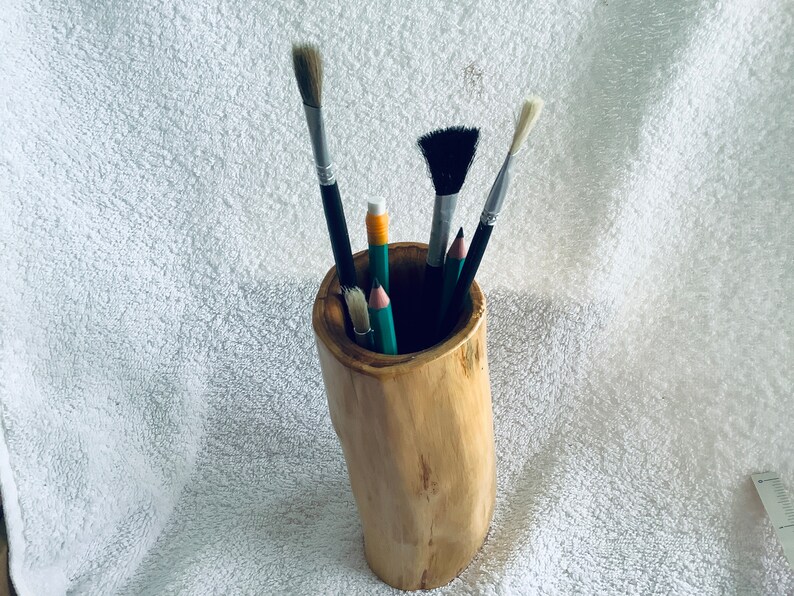 May include: A wooden pencil holder with a natural bark finish, filled with various paintbrushes and pencils. The holder is on a white fluffy surface.