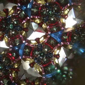 May include: A kaleidoscope image with a symmetrical pattern of red, blue, green, and gold. The image is made up of many small pieces of glass that are arranged in a geometric pattern.