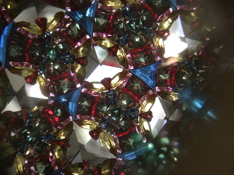 May include: A kaleidoscope image with a symmetrical pattern of red, blue, green, and gold. The image is made up of many small pieces of glass that are arranged in a geometric pattern.