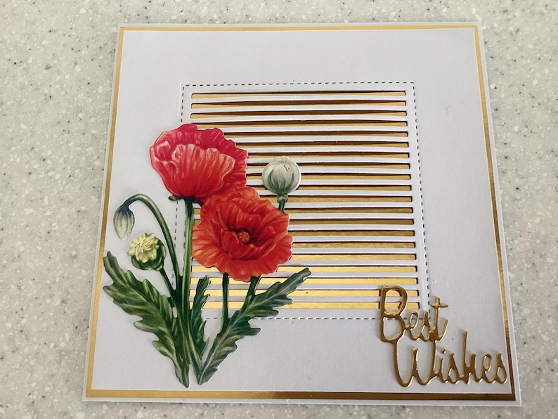 May include: A white greeting card with gold detailing. It features a floral design with red poppies and green leaves. The words "Best Wishes" are in gold script. The card is square in shape.