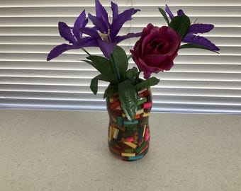 May include: A decorative vase filled with purple clematis and a deep red rose. The vase is made of a clear material and is decorated with colorful, rectangular pieces. The flowers and vase are set against a window with white blinds.