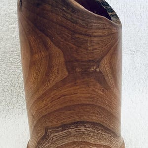 May include: A cylindrical wooden vessel with a natural wood grain pattern and a rough, dark bark-textured top edge. A yellow ruler is positioned behind the object, showing a height measurement of approximately 6 inches from the base to the top.