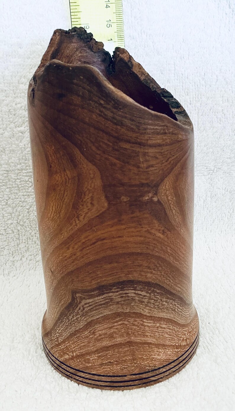 May include: A cylindrical wooden vessel with a natural wood grain pattern and a rough, dark bark-textured top edge. A yellow ruler is positioned behind the object, showing a height measurement of approximately 6 inches from the base to the top.