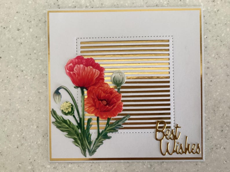 May include: A white greeting card with a gold border and accents. The card features a watercolour illustration of red poppies with green leaves. Gold horizontal stripes and the words "Best Wishes" are also included.