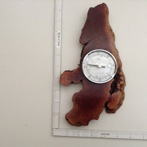 May include: A round, silver-coloured barometer with a white face and black markings is mounted on a brown, irregularly shaped piece of wood. The barometer is approximately 10 cm in diameter.