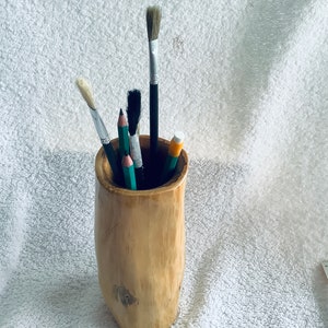 May include: A wooden pencil holder with a natural finish. It holds a variety of art supplies, including paintbrushes and pencils.