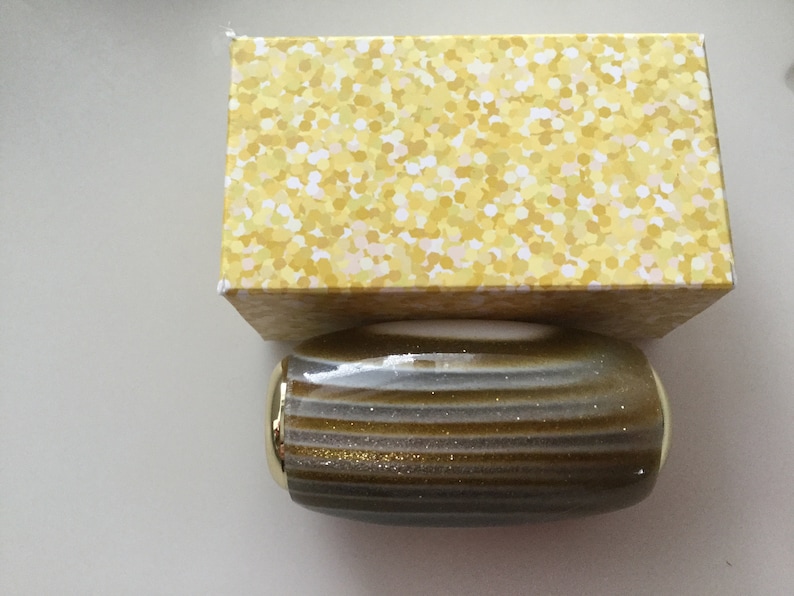May include: A gold and gray striped cylindrical container with gold accents. The container is sitting on top of a yellow box with a white polka dot pattern.