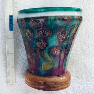 May include: A decorative, hand-painted vase with a wooden base. The vase is approximately 13 cm tall and features a teal, purple, and gold floral design. The rim is white and teal. The vase is a unique piece of home decor.
