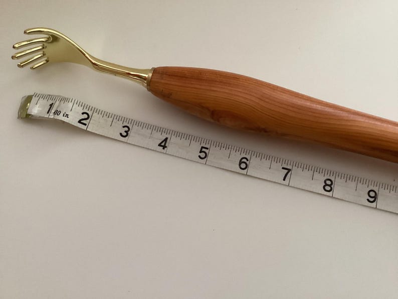 May include: A wooden and gold back scratcher with a long handle and a claw-like end. The scratcher is measured against a tape measure, showing it is approximately 18 cm long.