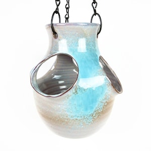 May include: A ceramic hanging planter with a light blue and brown glaze. The planter has three openings for plants and is suspended from a black chain.