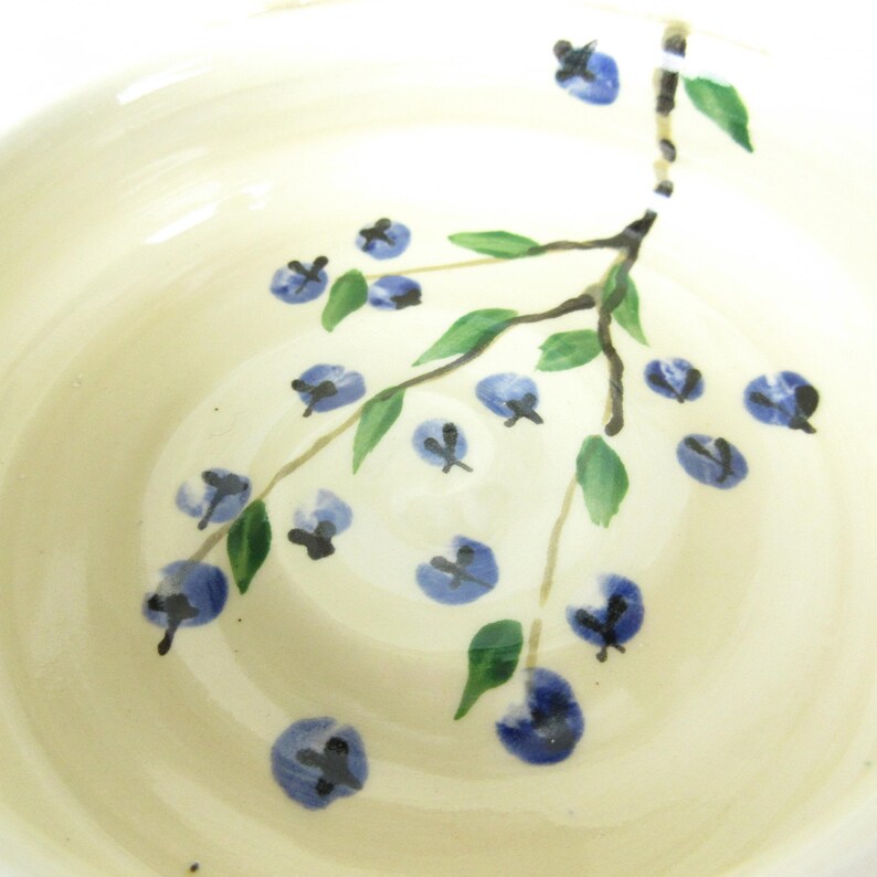 Puede incluir: Un taz&oacute;n de cer&aacute;mica blanco con un dise&ntilde;o floral azul y verde. El dise&ntilde;o presenta una rama con hojas y flores azules.