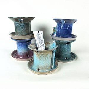 May include: A collection of handmade ceramic toothbrush holders in various colors, including blue, green, and brown. Each holder has a circular base and a cylindrical body with a hole for a toothbrush and toothpaste. One holder contains a tube of toothpaste and a toothbrush.