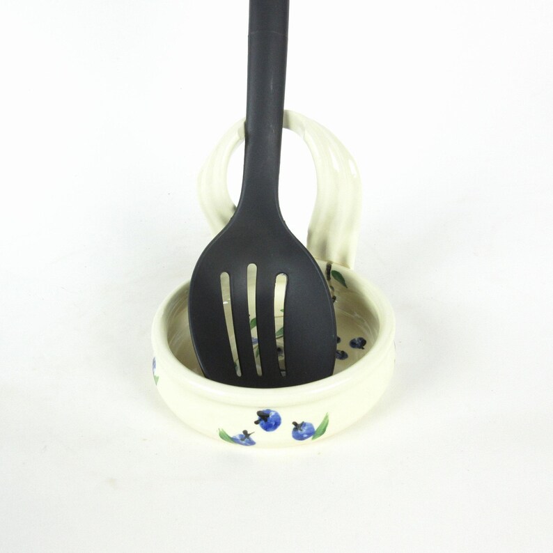 Puede incluir: Reposacucharas de cer&aacute;mica blanca con un dise&ntilde;o de ar&aacute;ndanos azul y verde. Una cuchara ranurada negra descansa en el soporte.