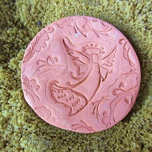 May include: A round, terracotta-colored ceramic piece featuring an embossed angel design. The angel has wings and a halo, surrounded by floral scrollwork. The piece is set against a background of small, golden-brown granules.