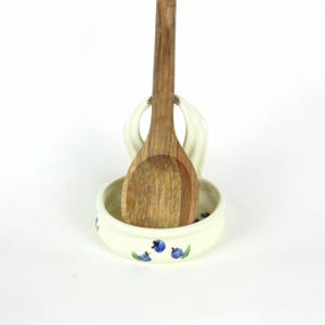 Puede incluir: Reposacucharas de cer&aacute;mica blanca con un dise&ntilde;o de ar&aacute;ndanos azul y verde. Una cuchara de madera descansa en el reposacucharas.