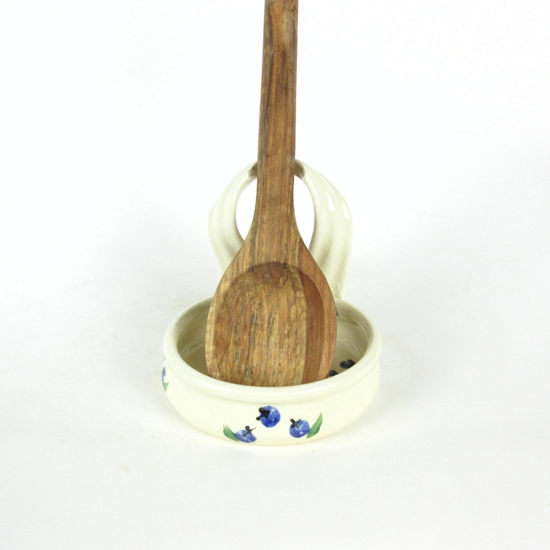 Puede incluir: Reposacucharas de cer&aacute;mica blanca con un dise&ntilde;o de ar&aacute;ndanos azul y verde. Una cuchara de madera descansa en el reposacucharas.