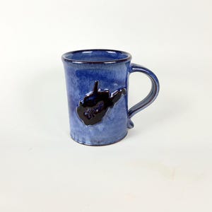 May include: A blue ceramic mug with a dark blue West Virginia state outline. The mug has a handle and a slightly textured surface. The mug is a deep blue color with a glossy finish.