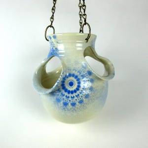 May include: A hanging ceramic planter with a cream-colored base and blue floral design. The planter has two large, curved handles and is suspended by a metal chain. The blue design is concentrated in the center and around the top rim.