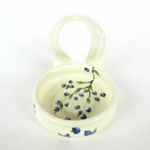 Puede incluir: Un plato de cer&aacute;mica blanco con asa, decorado con un dise&ntilde;o pintado a mano de ar&aacute;ndanos azules y hojas verdes.