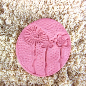 May include: A round, pink stamp with a floral design featuring three stylized flowers. The stamp has a textured surface and is set against a background of light brown granules. The flowers include a sunflower, a daisy, and a rose.