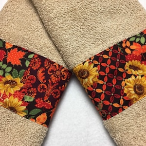 May include: Two tan hand towels with a decorative border of fall-themed fabric. The fabric features sunflowers, leaves, and a paisley pattern in shades of orange, red, brown, and green.