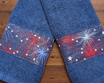 May include: Two blue hand towels with a decorative band featuring a fireworks design in red, white, and blue. The towels are made of a soft, absorbent material, perfect for the kitchen or bathroom.