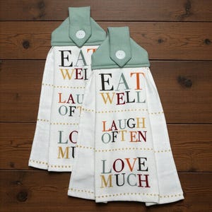 May include: Two white hanging kitchen towels with sage green fabric tops secured by white buttons. The towels feature the embroidered text EAT WELL, LAUGH OFTEN, and LOVE MUCH in alternating colors of dark gray, orange, maroon, and mustard yellow.