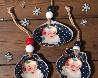 May include: Three seashell Christmas ornaments. Each shell is painted black with white polka dots and features a Santa Claus image. The ornaments are strung with twine and wooden beads in red, white, and black.