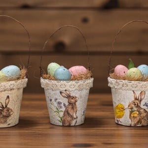 May include: Five decorative Easter baskets with wire handles. Each basket is filled with faux straw and colorful eggs. The baskets feature vintage-style illustrations of bunnies and chicks, with lace trim around the top.