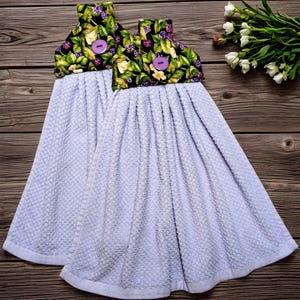 May include: Two light blue kitchen hand towels with a decorative top featuring a black background with green leaves, purple berries, and yellow flowers. Each towel has a purple button. The towels are hanging on a wooden surface.