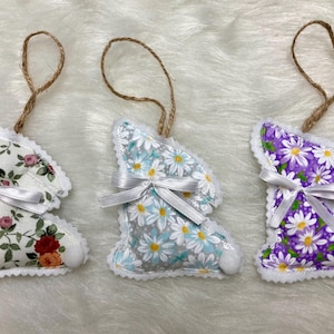 May include: Three handmade fabric bunny ornaments with white lace trim. The bunnies are decorated with floral patterns in shades of purple, blue, and white.
