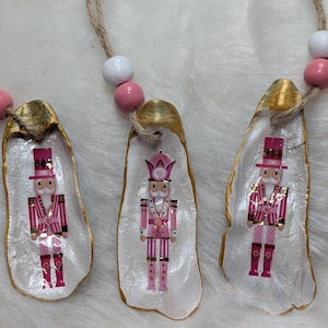 May include: Three Christmas ornaments featuring pink and white nutcracker designs on oyster shells. The shells have gold-painted edges and are strung with pink and white beads. The ornaments are on a white, textured surface.