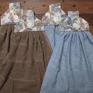 May include: Four hanging kitchen towels. Two are brown, and two are blue. Each towel has a patterned top with paisley designs in shades of blue, brown, and cream. The tops are secured with a white button.