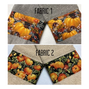 May include: Two kitchen towels with a fall theme. The top towel is gray with a brown and orange pumpkin print. The bottom towel is beige with a black and orange pumpkin print. The text "FABRIC 1" and "FABRIC 2" is printed on the towels.